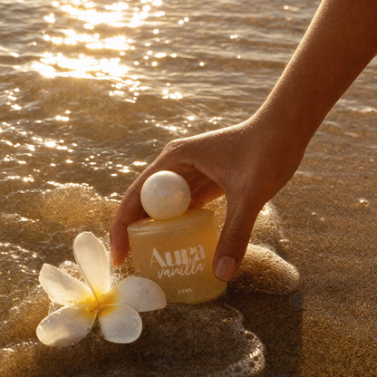 Main attrapant le flacon Aura Vanilla posé sur le sable à côté d'une fleur de frangipanier