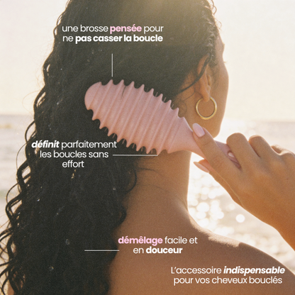 Femme démêlant ses cheveux bouclés avec la brosse Sculptor rose au bord de la mer